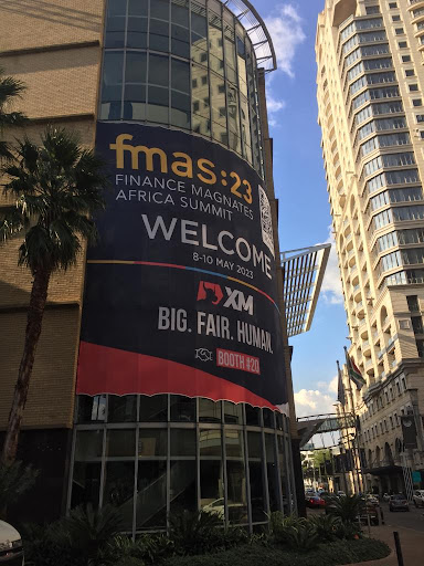 An event banner displayed across the side of a building, reading “FMAS23 Finance Magnates Africa Summit Welcome 8 10th May 2023” with the XM logo followed by “Big Fair Human Booth #20”